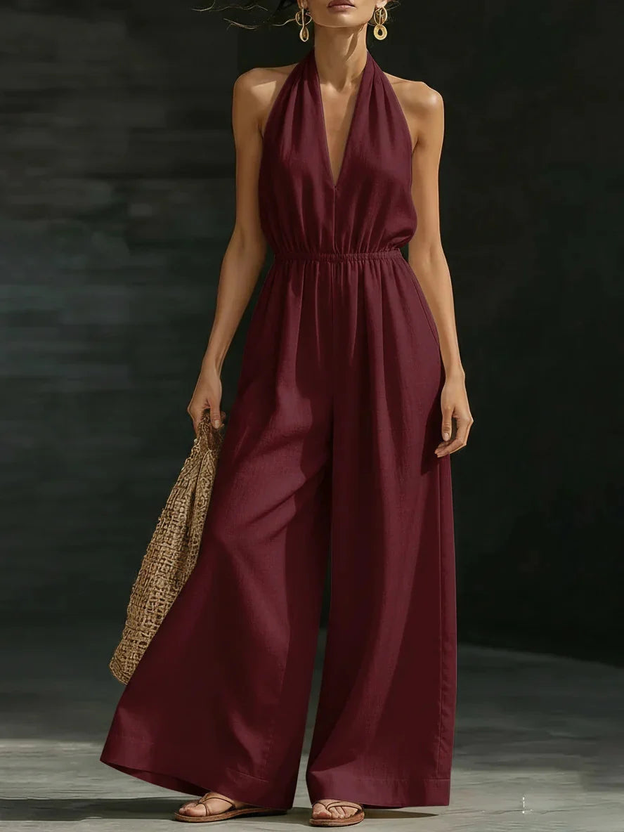 Woman in deep burgundy jumpsuit with halter neck and wide legs, holding woven bag in dim studio setting.