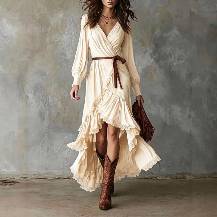 Woman in cream wrap dress with long sleeves, lace trim, layered skirt. Holding brown fringe bag, wearing cowboy boots, gray backdrop.