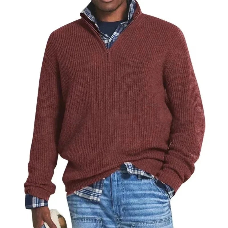 Man wearing a red zip sweater over a plaid shirt with jeans, holding a hat in a studio setting.