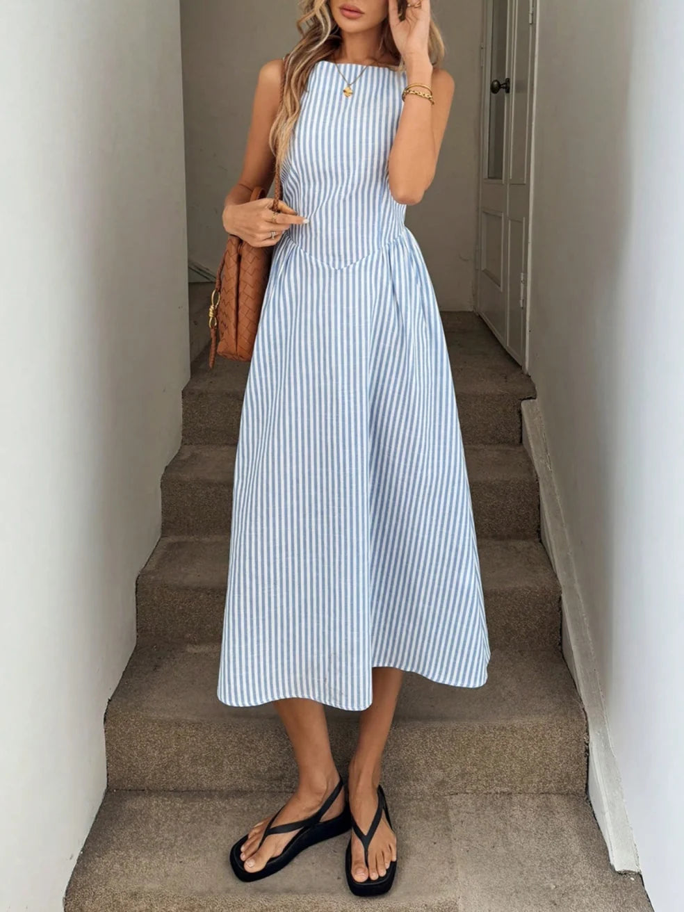 Woman wearing a blue and white striped maxi dress with a sleeveless design, standing on indoor steps with a bag.