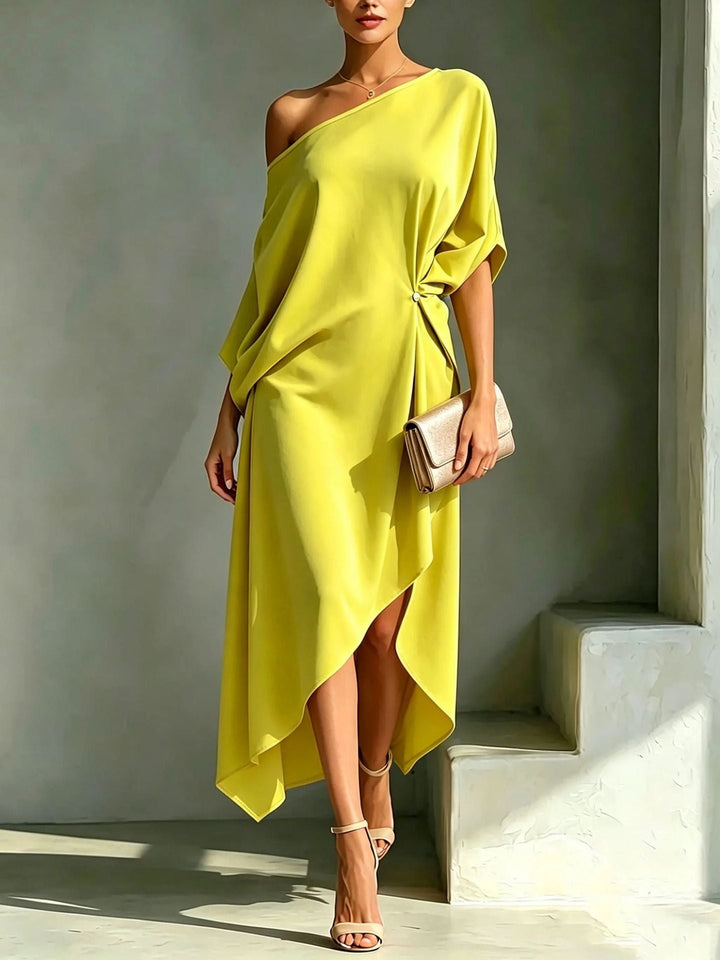 Elegant asymmetrical yellow robe with off-shoulder design, styled with a clutch and strappy sandals in a minimal studio setting.
