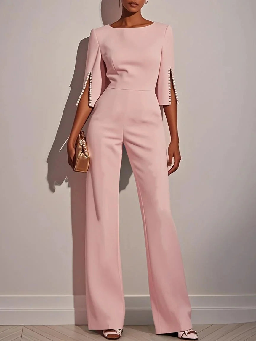 Woman in a pink jumpsuit with pearl-adorned sleeves holds a gold clutch in a minimalist indoor setting.
