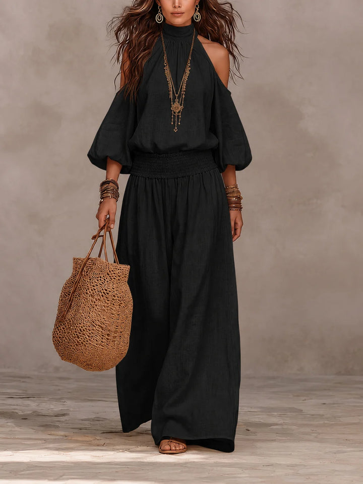Model in black jumpsuit featuring off-shoulder style, paired with jewelry and a woven bag, set against a minimalist backdrop.