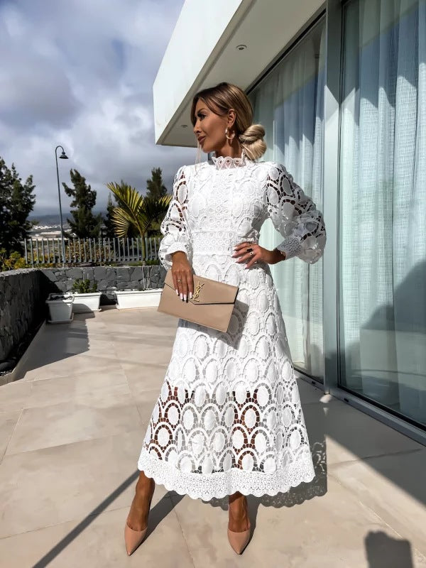 White lace dress in an outdoor setting, accessorized with a beige clutch and nude heels, posing near a window with trees visible.