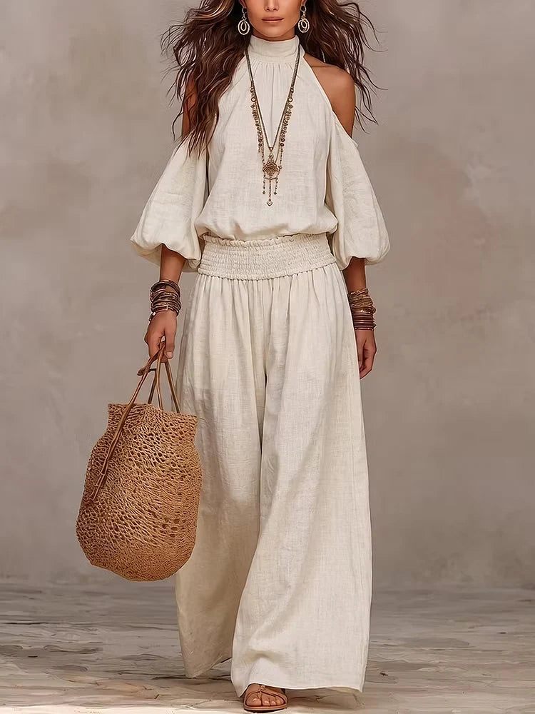 Model showcasing cream-colored off-shoulder jumpsuit with jewelry and a woven bag, in a subtle studio environment.