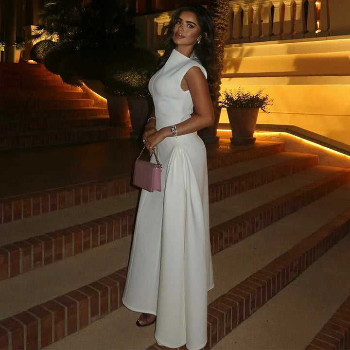 Side view of woman in a white elegant dress on brick steps at night, holding a pink handbag with warm lights behind.