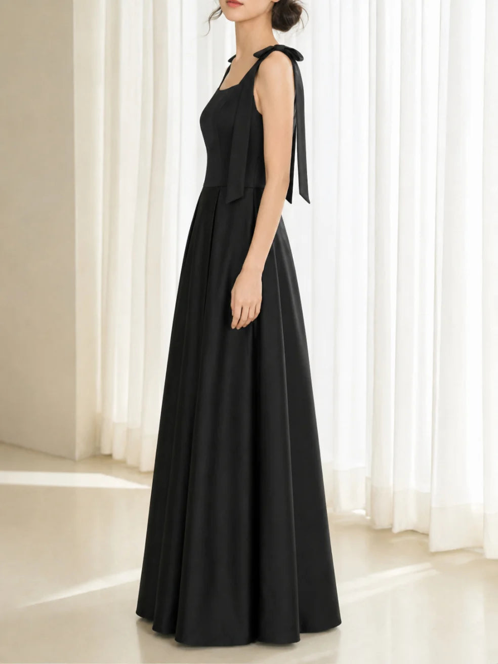 Side view of an elegant black dress with bow straps in a well-lit interior with sheer curtains.