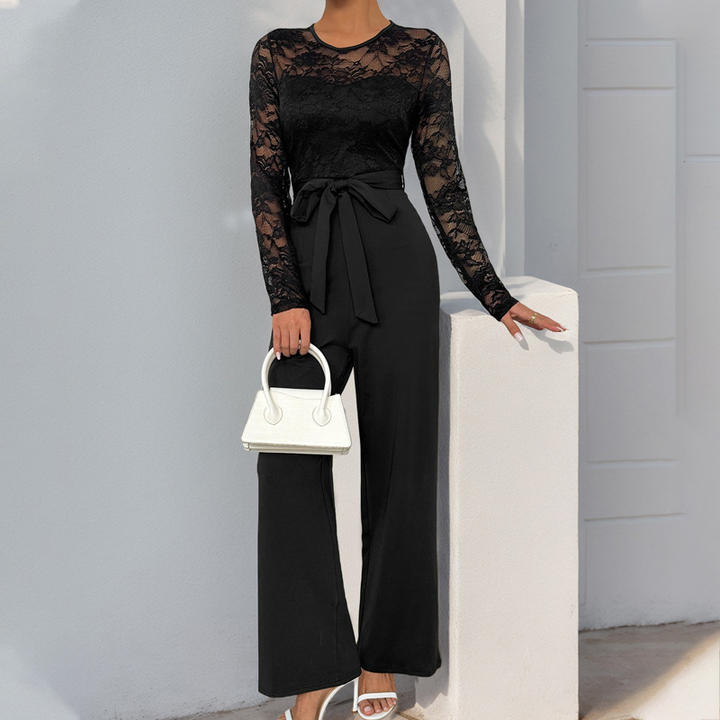 Black lace bell sleeve jumpsuit with a waist tie, paired with a white handbag and sandals, outdoor light setting.