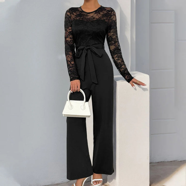Sophisticated black jumpsuit featuring lace sleeves and a waist tie, accessorized with a white handbag, outdoors by a light wall.
