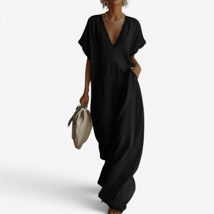 Black maxi dress with lace-trimmed V-neckline, worn by a woman holding a beige bag, standing against a plain background.