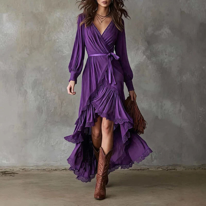 Woman in purple wrap dress with long sleeves, lace trim, layered skirt. Holding brown fringe bag, wearing cowboy boots, gray backdrop.