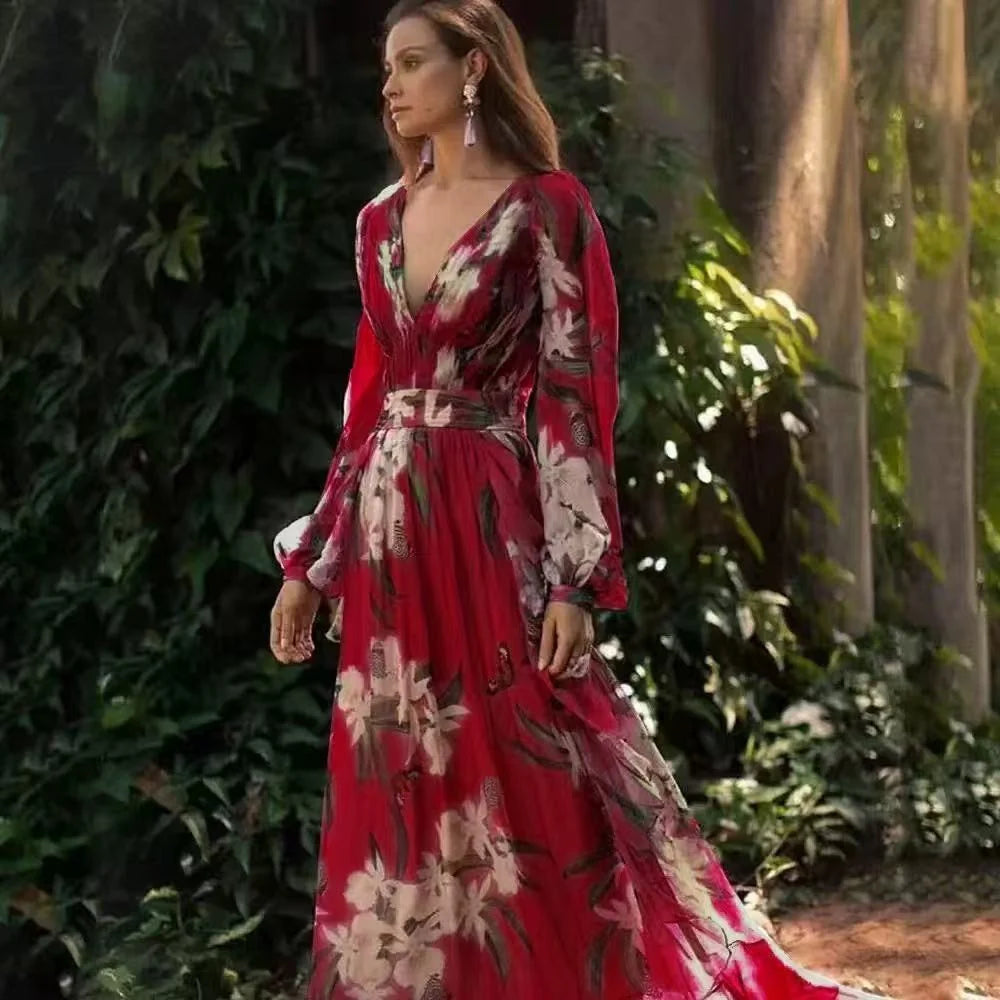 Woman in a flowing red floral maxi dress with long sleeves, standing outdoors by lush greenery.