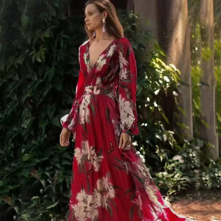 Woman in a flowing red floral maxi dress with long sleeves, standing outdoors by lush greenery.