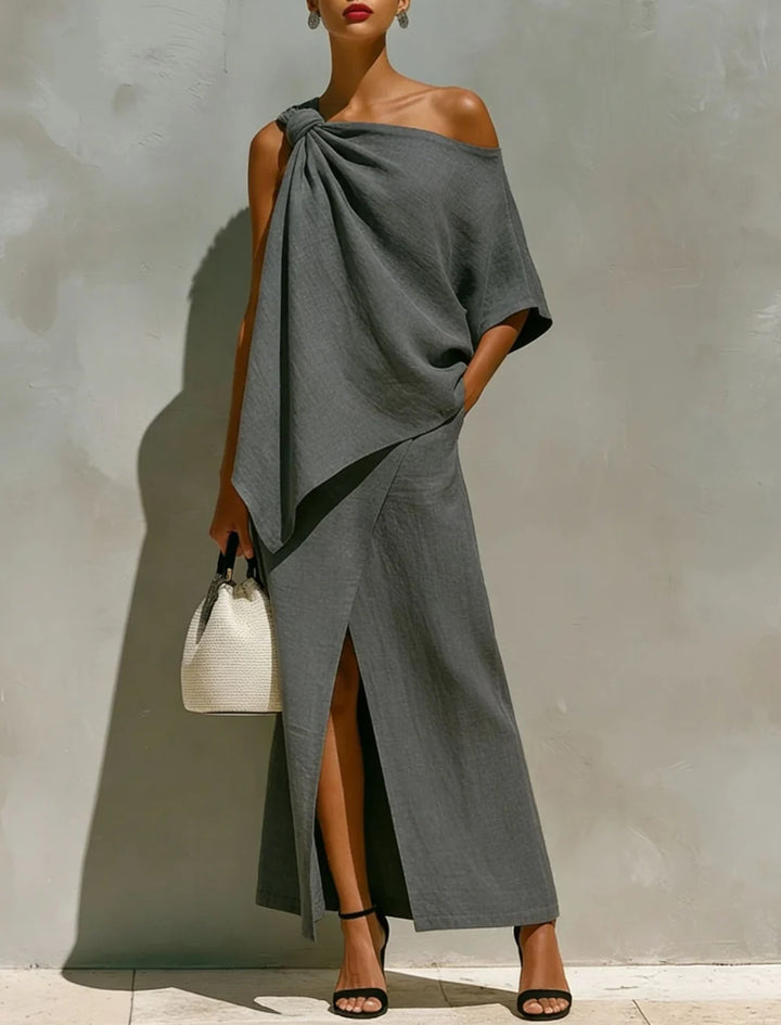 Chic gray two-piece set with an off-shoulder top and long skirt with a slit, displayed in a well-lit studio, complemented by black heels and a woven bag.