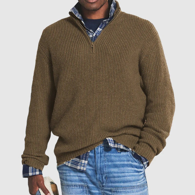 Man wearing a brown zip sweater over a plaid shirt with jeans, holding a hat in a studio setting.