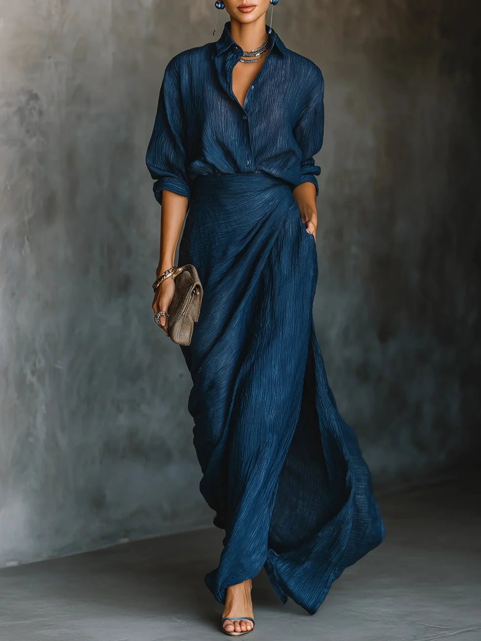 Woman wearing a flowing, blue maxi dress with rolled sleeves, holding a clutch in an indoor setting with a textured gray wall.
