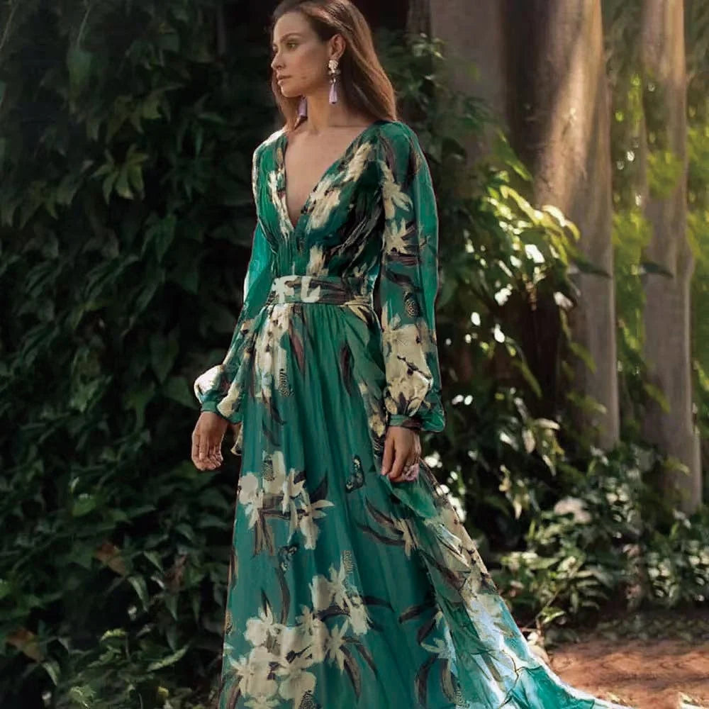 Woman in a flowing green floral maxi dress with long sleeves, standing outdoors by lush greenery.