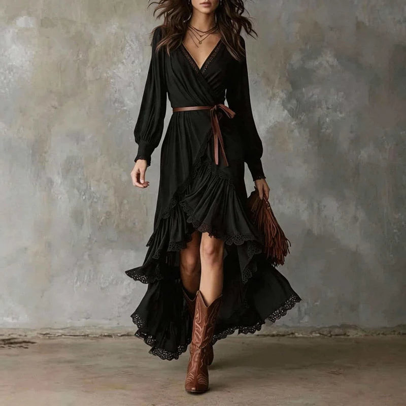 Woman in black wrap dress with long sleeves, lace trim, layered skirt. Holding brown fringe bag, wearing cowboy boots, gray backdrop.
