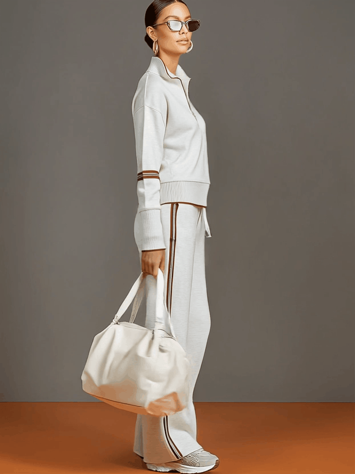 Side view of a matching lounge set in light gray with contrast stripes, styled with white sneakers and a bag in a studio setting.