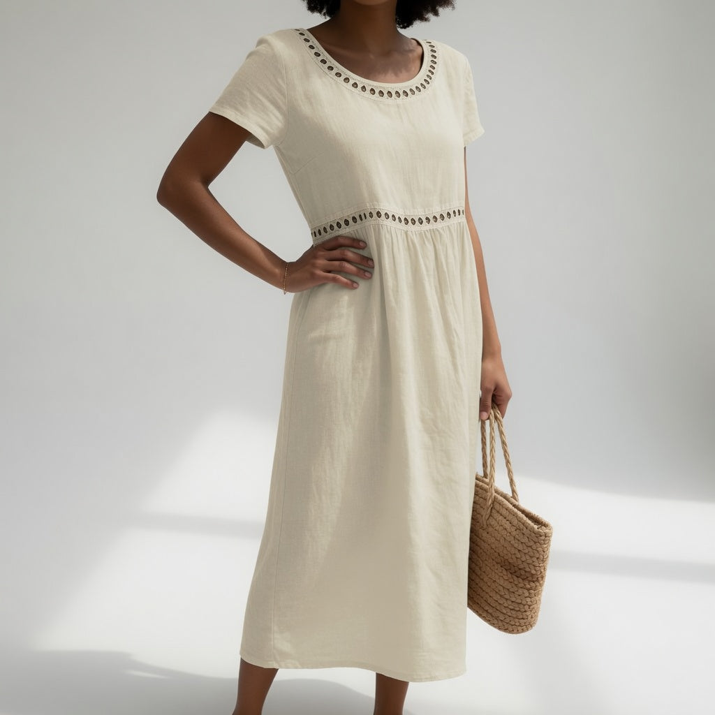 Cream midi dress with eyelet accents and short sleeves, modeled by a person with a woven bag. Simple studio background.