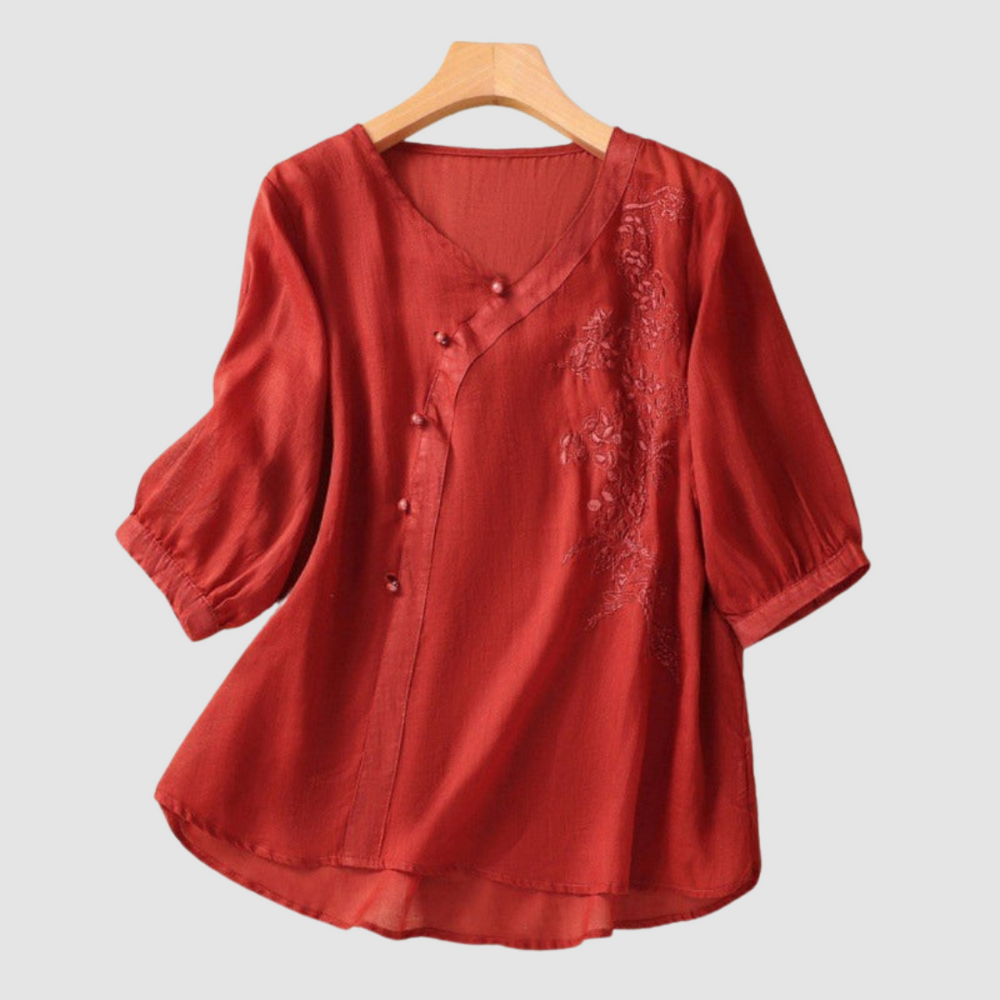 Rust-colored blouse with floral embroidery and subtle buttons on hanger against white background.