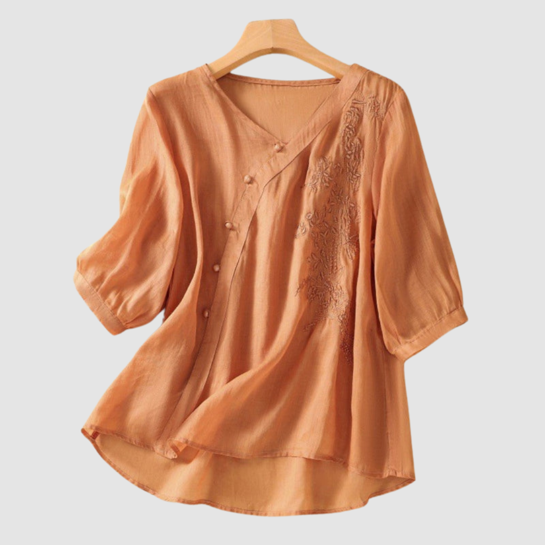 Soft peach blouse with floral embroidery and delicate buttons on hanger against white background.