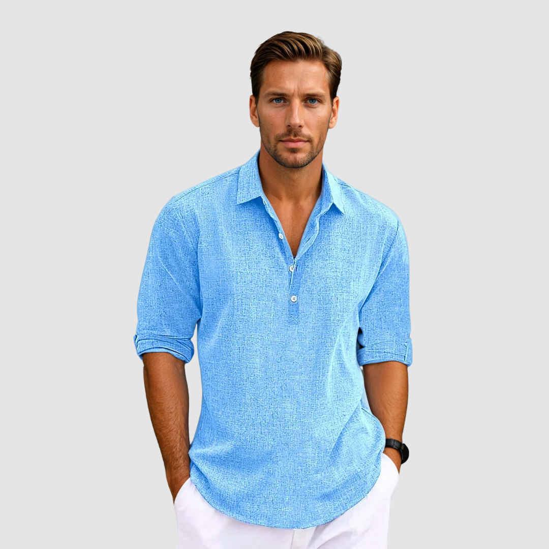 Man wearing a blue collared shirt with rolled up sleeves, holding hands in pockets, against a light gray background.