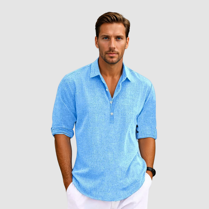 Man wearing a blue collared shirt with rolled up sleeves, holding hands in pockets, against a light gray background.