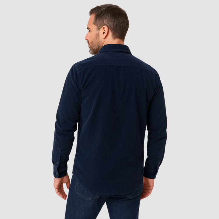 Rear view of a man wearing a navy blue corduroy shirt and jeans, standing against a plain light gray background.