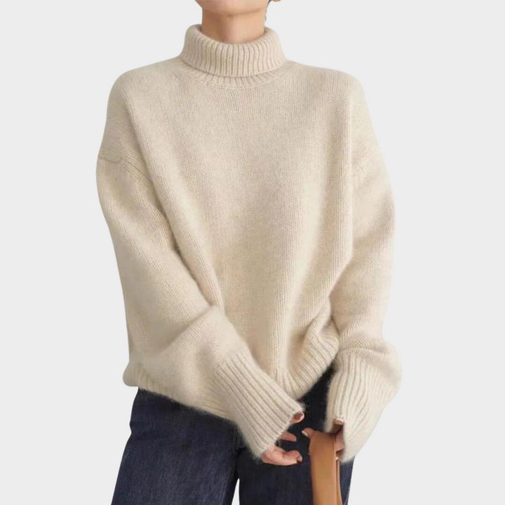 Beige soft turtleneck jumper worn by a model in blue jeans, holding a brown handbag, studio background.