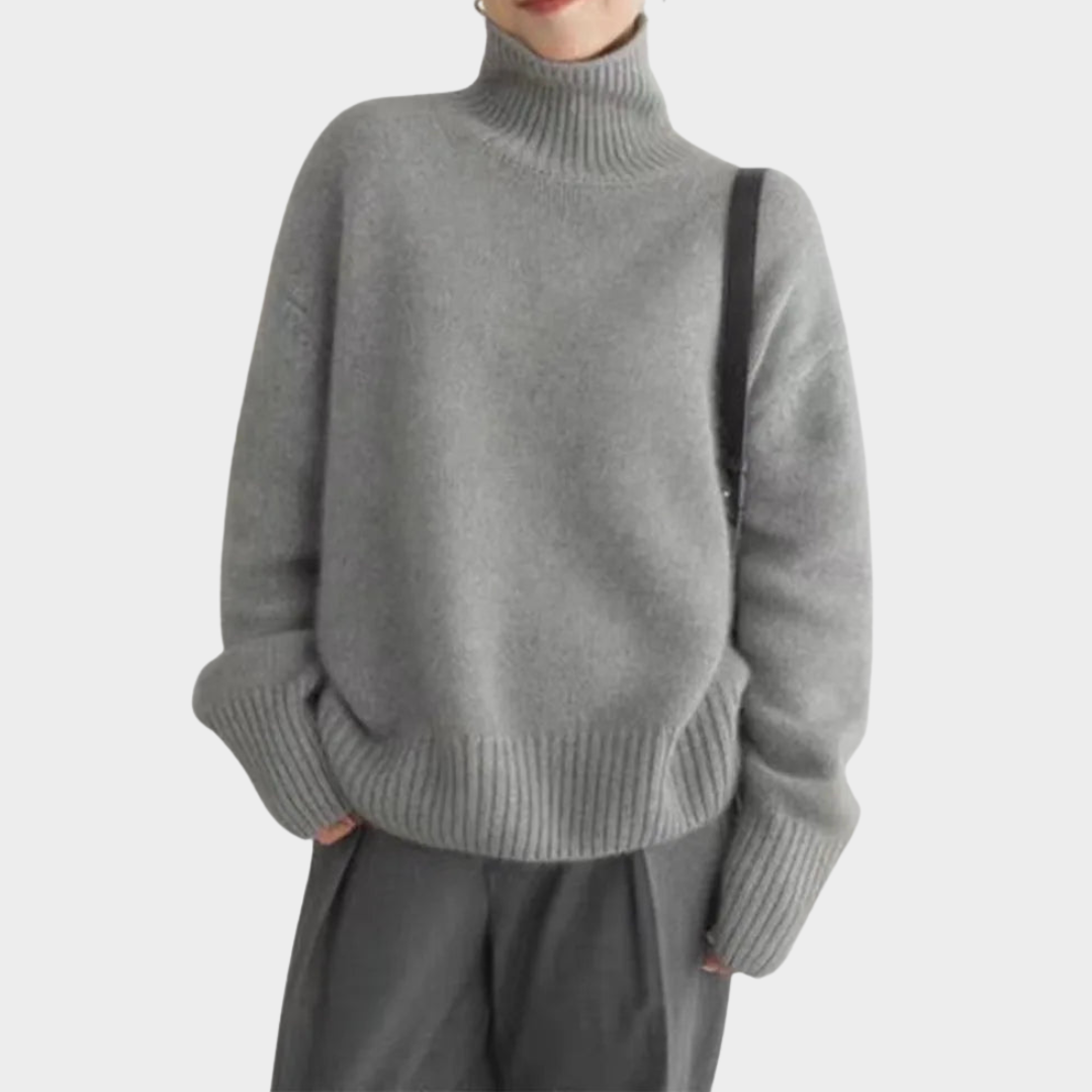 Gray soft turtleneck jumper worn by a model with gray pants and a black shoulder strap, studio background.