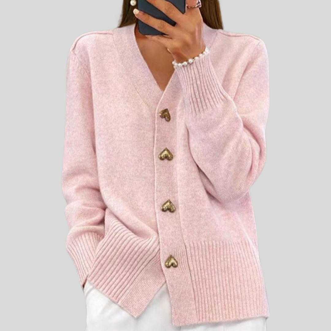 Model wearing a soft pink cardigan with heart-shaped buttons, paired with white pants against a gray background.