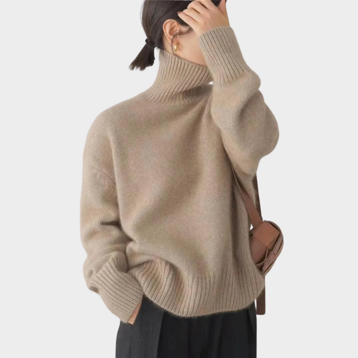 Taupe soft turtleneck jumper with ribbed accents, worn by a model with black pants and a brown crossbody bag, studio background.