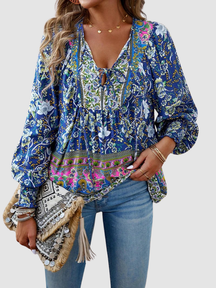 Vibrant blue floral blouse with colorful details, styled with light jeans and a decorative clutch, against gray backdrop.