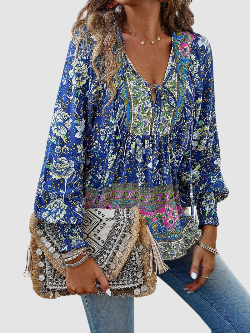 Front view of blue floral blouse with elaborate patterns, paired with jeans and a textured clutch, on gray background.