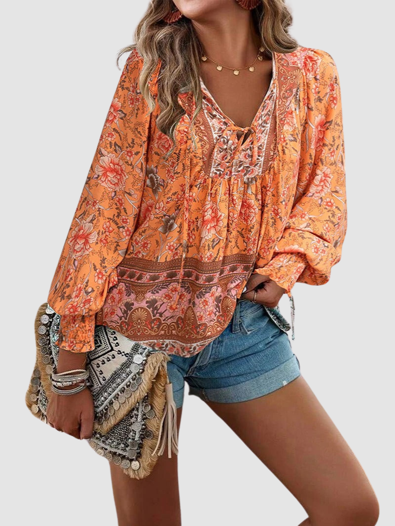 Front view of orange floral blouse with ornate patterns, worn with denim shorts and a decorative clutch, on neutral background.
