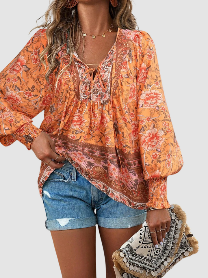 Bright orange floral blouse with intricate designs, paired with denim shorts and a detailed clutch, on gray backdrop.