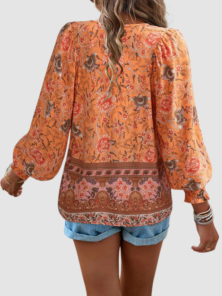 Back view of orange floral blouse with detailed designs, styled with denim shorts, against a gray backdrop.
