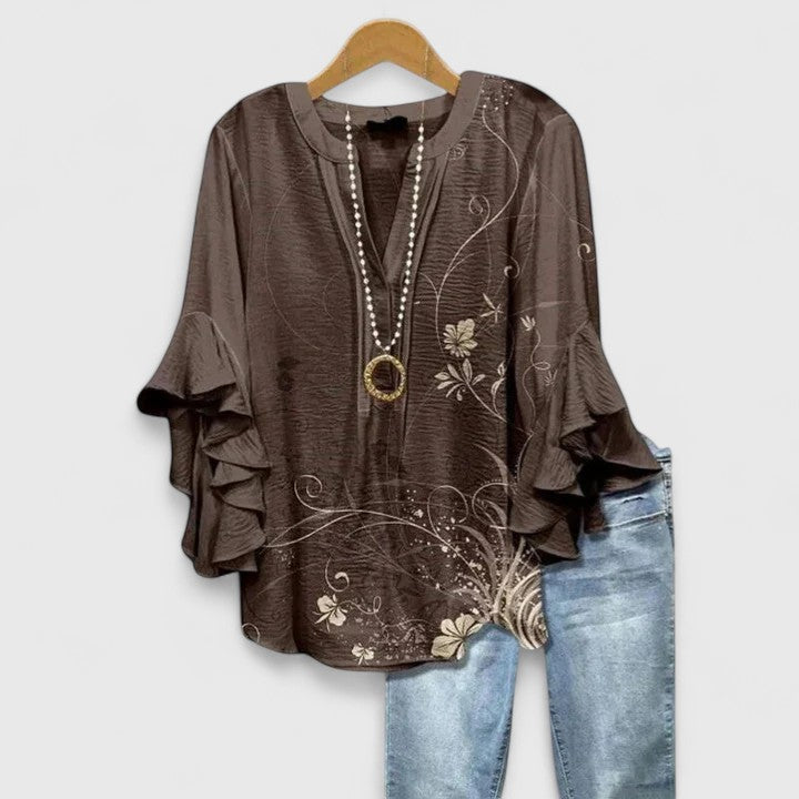 Dark brown blouse with beige floral accents and ruffle sleeves, paired with light jeans, on wooden hanger.