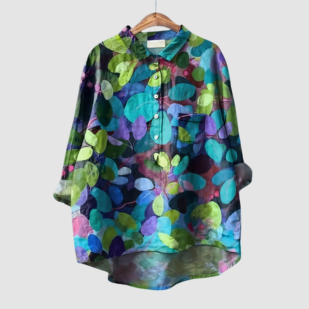 Colorful leaf pattern blouse with vibrant greens and purples on wooden hanger against light gray background.