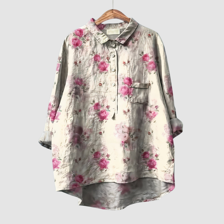 Soft pink floral blouse on wooden hanger with subtle background detailing against light gray background.