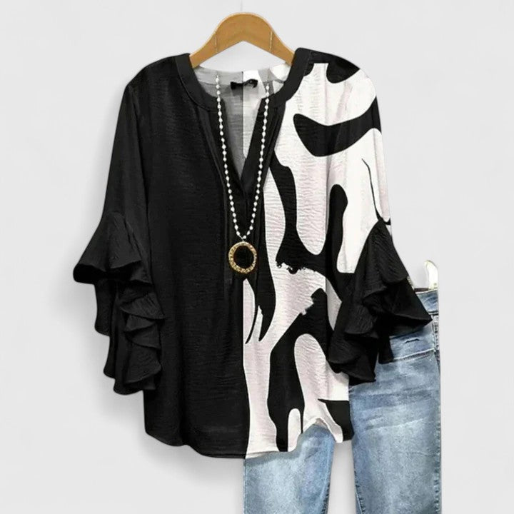 Black and white abstract patterned blouse with ruffle sleeves, alongside light jeans, on wooden hanger.