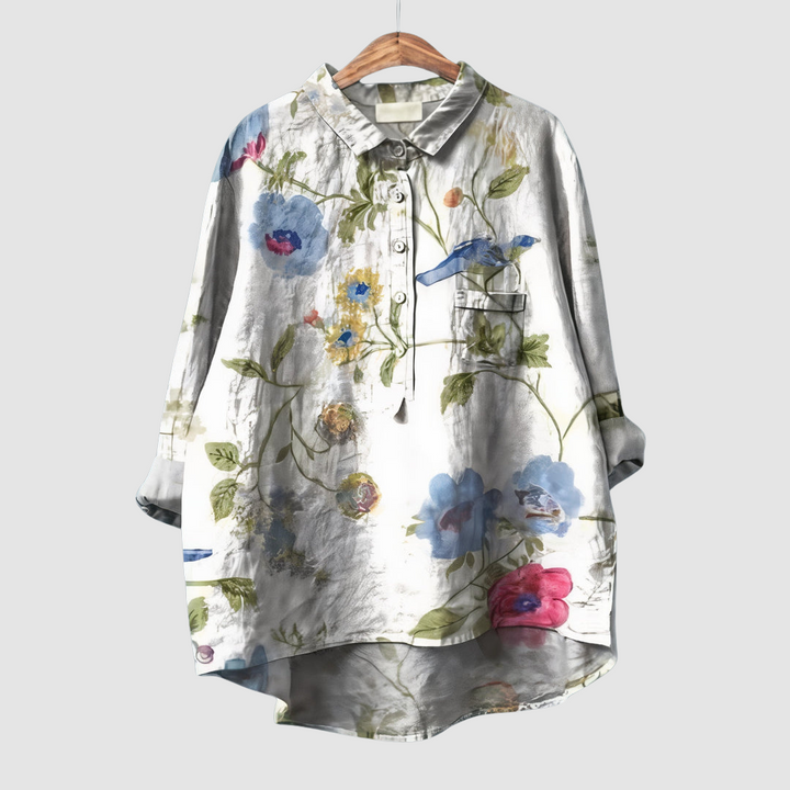 Light colored blouse with subtle floral and bird print on wooden hanger against light gray background.