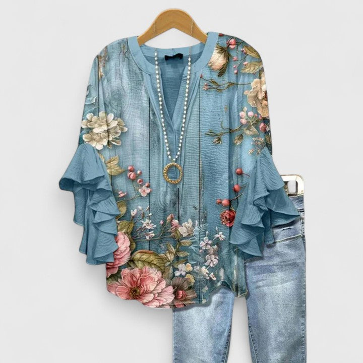 Blue floral blouse with vibrant flower motifs and ruffle sleeves, complemented by light jeans, on wooden hanger.