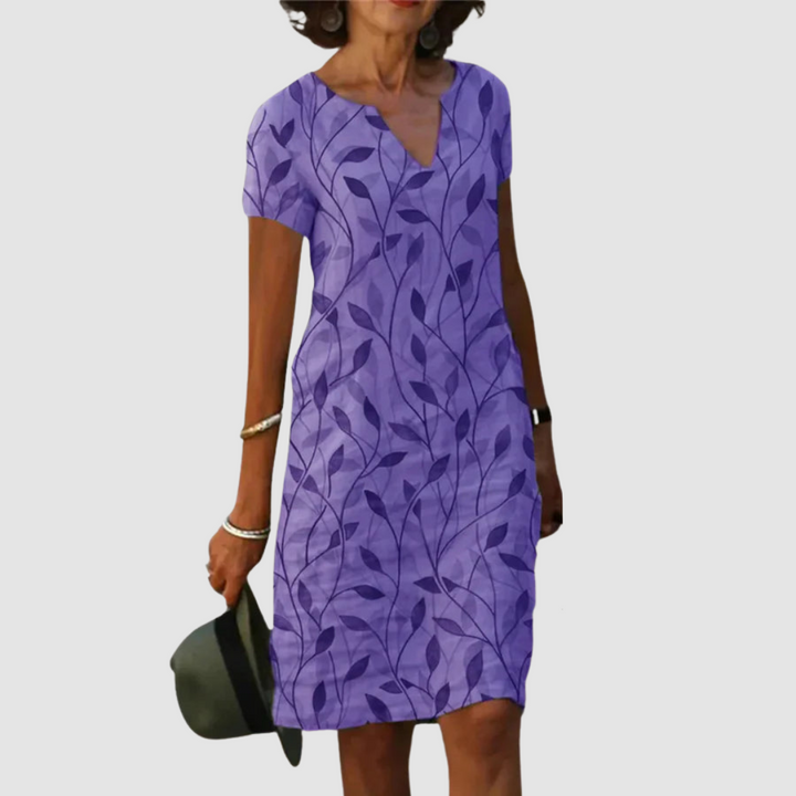 Model wearing a short-sleeve, purple dress with leaf print, holding a hat against a plain background.