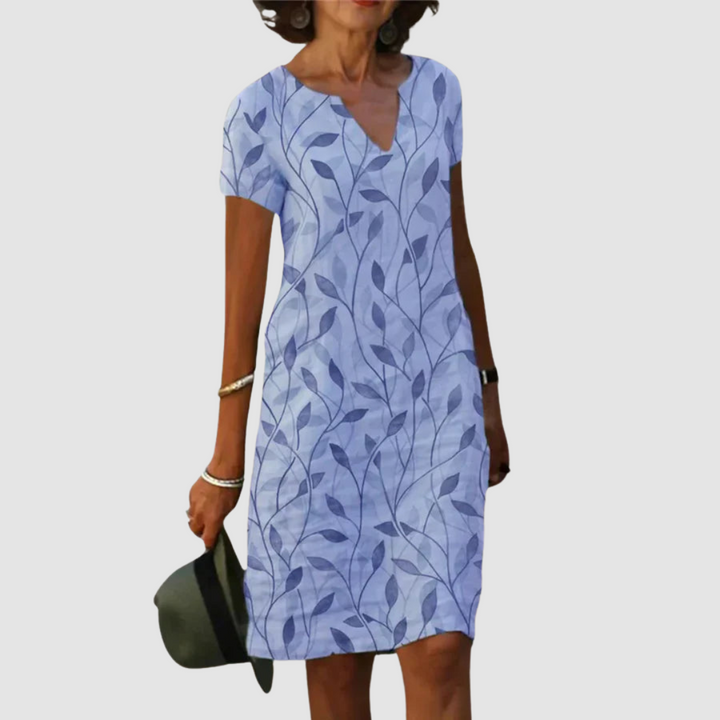 Model wearing a short-sleeve, light blue dress with leaf print, holding a hat against a plain background.