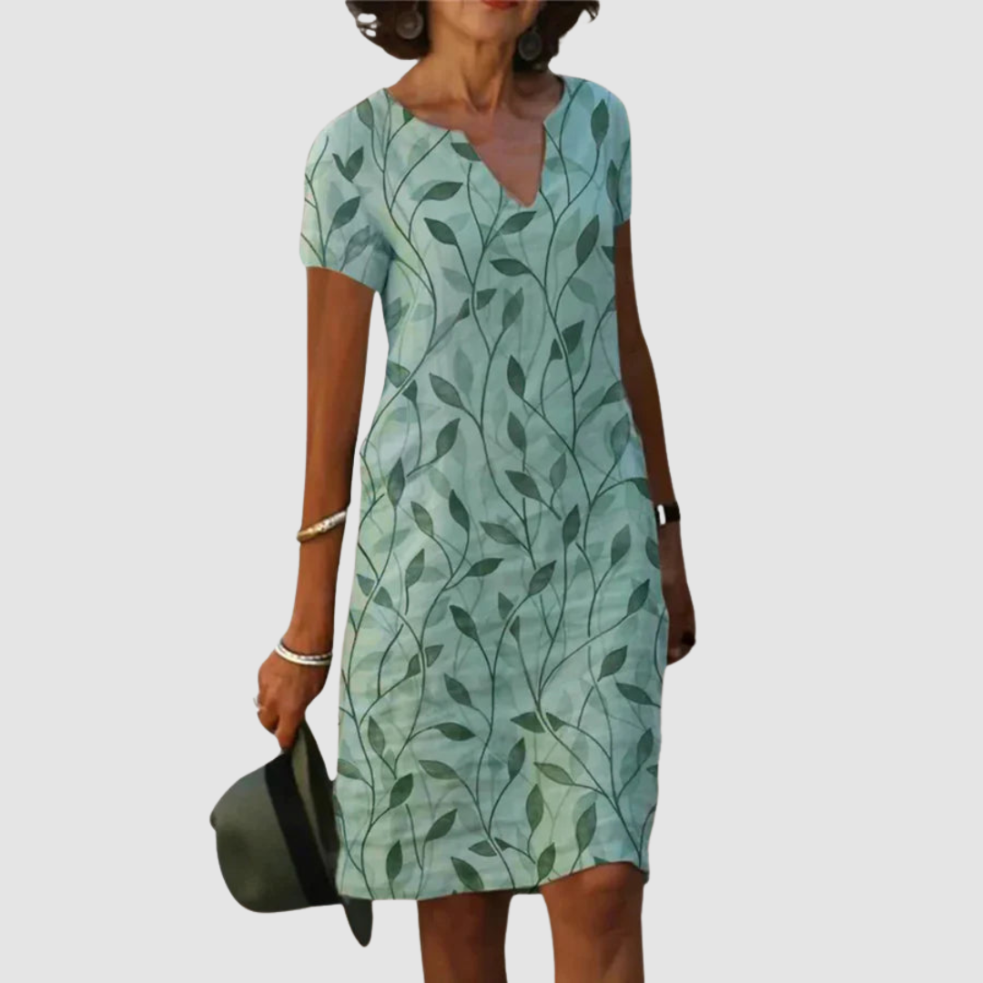 Model wearing a short-sleeve, light green dress with leaf print, holding a hat against a plain background.