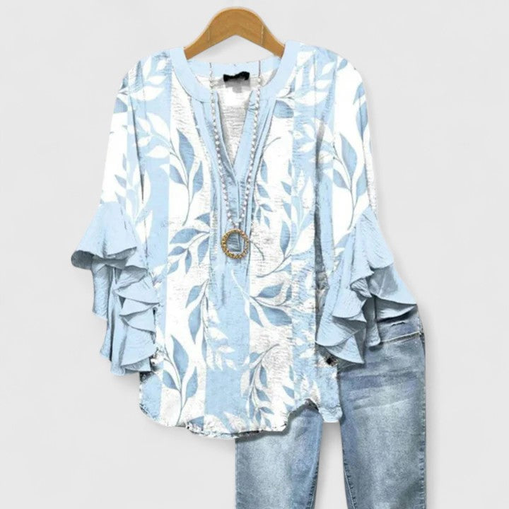 Light blue and white leaf patterned blouse with ruffle sleeves, with light jeans, on wooden hanger.