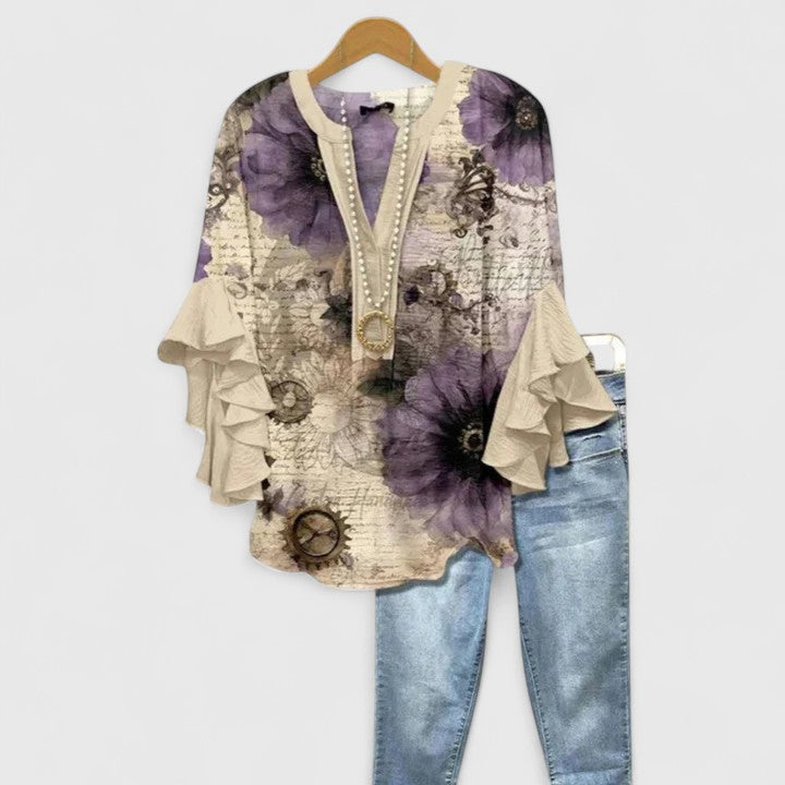 Beige blouse with purple floral design and ruffle sleeves, alongside light jeans, on wooden hanger.