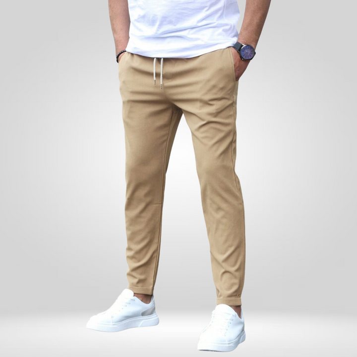 Tan stretch pants with drawstring, modeled with white t-shirt and sneakers on a gray studio backdrop.